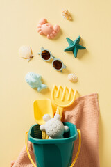 Flat lay of childrens beach toys including a green bucket with playful figures, yellow spade, rake on a soft pink towel against a sunny yellow backdrop. © photoguns