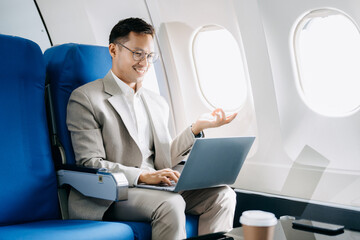 Fototapeta premium Attractive Asian male passenger of airplane sitting in comfortable seat while working laptop and tablet with mock up area using wireless connection.