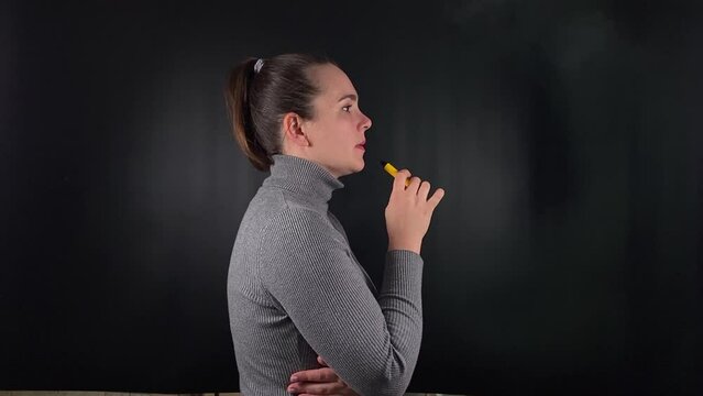 A charming young woman exhales a thick cloud of vapor, e-cigarette. With a casual, unbothered expression set on the stark black backdrop, youthful indulgence and the cavalier embrace of trending habit