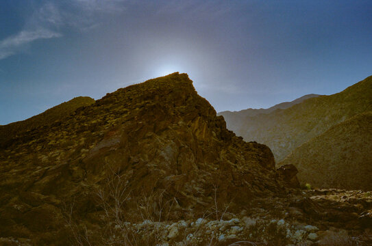 The sun hovers over the peak of a rugged mountain, casting a warm glow on the rocky landscape while the clear sky fades into a soft gradient.