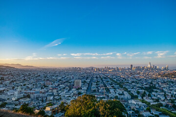 An aerial view showcases a dense urban cityscape at dusk, with rows of buildings leading to a skyline punctuated by skyscrapers under a clear blue sky.