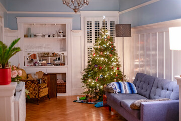 A cozy living room decorated for Christmas, featuring a lit Christmas tree, a grey sofa, and a kitchen in the background.