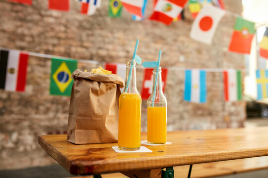A table with two bottles of orange juice and a brown paper takeout bag against a backdrop of international flags.