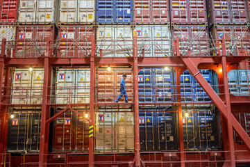 A worker is walking along the narrow gangway of a large cargo ship, surrounded by numerous colorful stacked shipping containers.