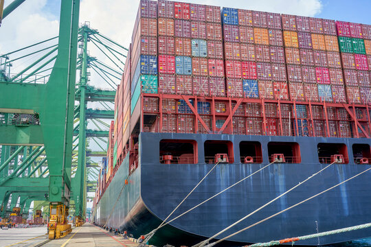 A large cargo ship loaded with colorful containers is docked at a port with cranes in the background.