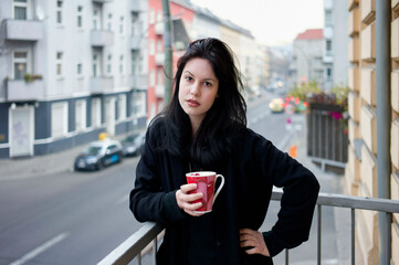 A woman stands on a balcony holding a red mug, with a street scene in the background.