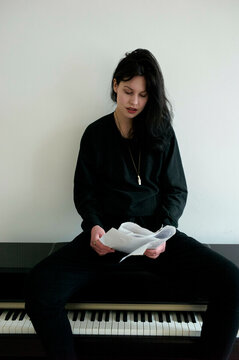 A young woman sits on a piano bench, dressed in black attire, perusing sheets of music with a thoughtful expression.