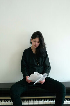 A woman sits in front of a piano, reading a sheet of paper with a focused expression.