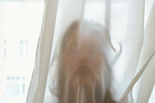 A person's silhouette is visible behind a sheer curtain, with their facial features slightly obscured and fingertips pressing against the fabric.