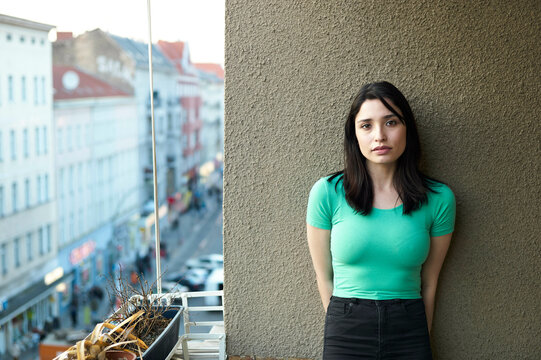 A woman in a green top stands against a wall on a balcony overlooking a city street.
