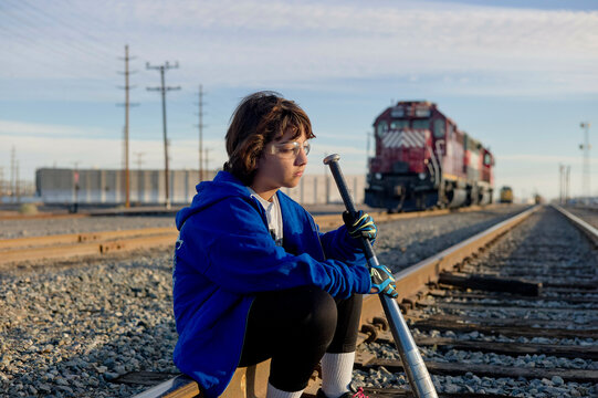 A person sits on a railroad track, holding a long pole, with a train and power lines in the background during the daytime.