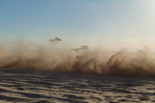 A vehicle speeds across the desert, kicking up a massive cloud of dust in the wake of its passage.