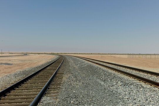 A view of railroad tracks extending straight into the horizon in a desert landscape under a clear blue sky.