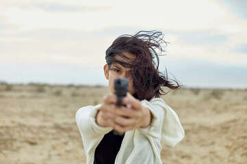 A person is pointing a gun directly at the camera with intense focus. Their hair is windswept, and they are dressed in a white jacket over a black top, set against a desolate landscape.