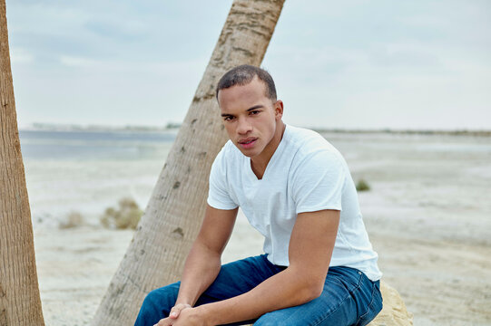 A man sits crouched by a tree trunk on sandy ground, gazing at the camera with a serene expression, with a clear sky and flat horizon in the background.