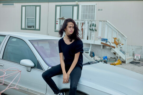 A woman sits on the hood of an old silver car in front of a building with a staircase and various items strewn around.
