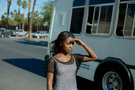 A young woman shields her eyes from the sun while walking on a sunny street with a bus and palm trees in the background.
