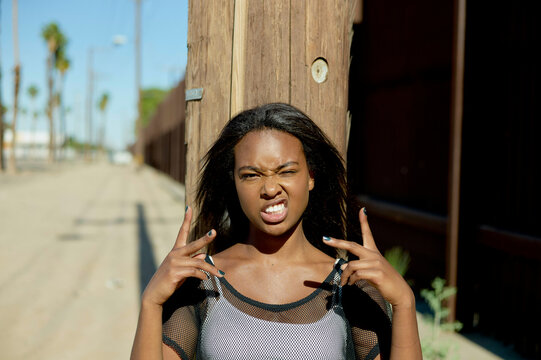 A young woman stands outdoors flashing a playful peace sign on each hand beside her face, with a slight frown, against a blurred background of palm trees and a wooden pole.