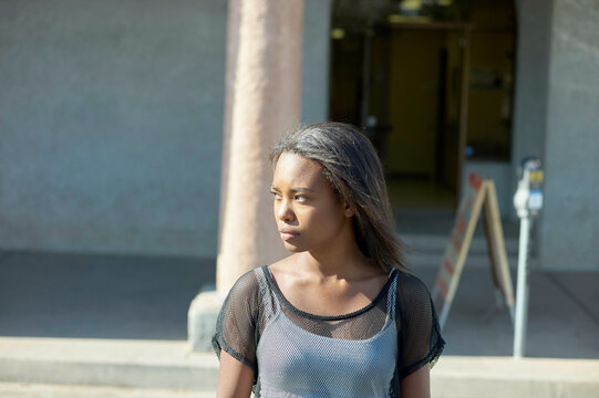 A young woman stands outdoors, gazing into the distance, her hair flowing over her shoulders, with a building entrance behind her.