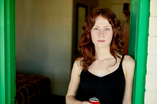 A red-haired woman is standing in a doorway with bright green trim, holding a drink can and gazing directly at the camera.