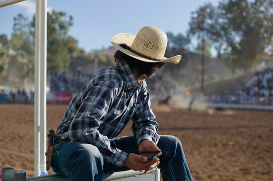 A person is sitting on a rail, wearing a cowboy hat and plaid shirt, looking at a mobile phone with a rodeo arena in the background.