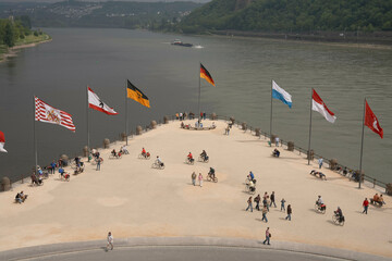 The image shows a scenic viewpoint with people walking and sitting alongside a river. National flags flutter on poles above the promenade against a backdrop of hills and water.
