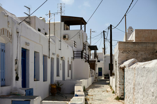 A narrow alley in a traditional Mediterranean village with white buildings and blue doors under a clear sky, with electrical wires crisscrossing overhead.