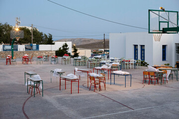 An outdoor setting at dusk featuring a basketball court with tables and chairs neatly arranged across it, under the glow of gentle lights, adjacent to a white building with blue trimmings.