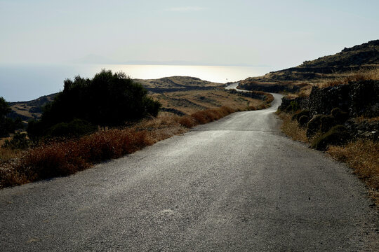 A winding road descends through a dry landscape towards the sea, flanked by shrubs and rocks under a hazy sky.