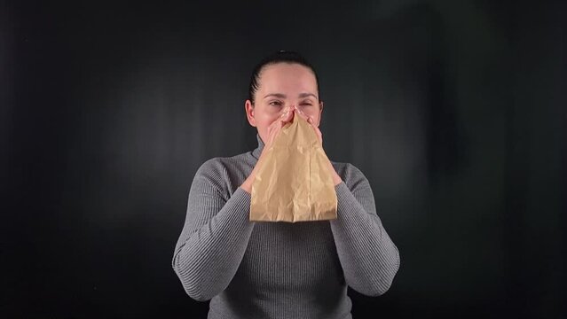 Portrait of a young woman breathing into a paper bag to calm herself during a panic attack on a black background. The struggle with anxiety and the methods used to find relief in moments of distress