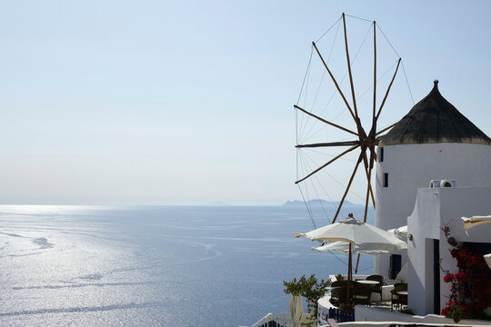 A traditional white windmill with a wooden windwheel overlooks the sparkling blue sea from a high vantage point, with a clear sky above and a hint of a distant island on the horizon.