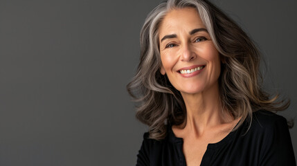 Fototapeta premium Beautiful face of a smiling mature woman glowing with the effects of shining anti-aging cosmetics, on a grey studio background, radiating confidence and happiness, highlighting sel