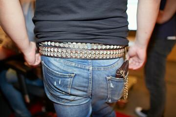 A close-up view of a person wearing blue denim jeans and a black shirt tucked in, with a studded belt and keychain accessory attached to a belt loop.