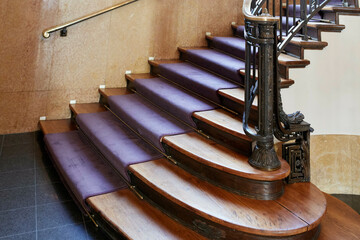 A polished wooden spiral staircase with purple carpeted steps, featuring a metal handrail and ornate banister.