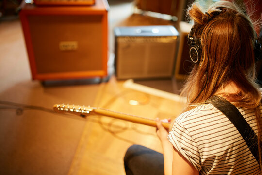 A person with blonde hair is sitting while playing an electric guitar, facing amplifiers in a room with warm lighting.