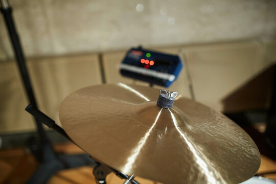 A close-up of a shiny cymbal as part of a drum set with a music amplifier lit by red LEDs in the soft focus background.