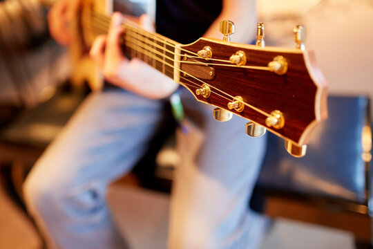 A close-up of a guitar headstock being played, with focus on the tuning pegs and blurred hands and strings in the background.