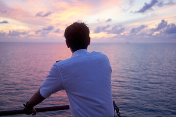 A man in a white shirt gazes out at the ocean from a ship's railing as the sky displays a beautiful gradient of sunset colors.