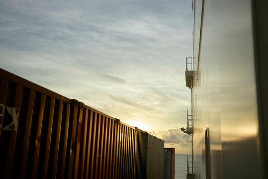 The image captures the side of a cargo ship with stacked shipping containers at sunset, with the sun dipping near the horizon, casting warm light and soft shadows.