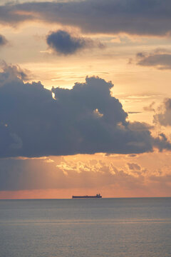 A cargo ship is silhouetted against the vibrant orange hues of a sunset sky, with large clouds partially obscuring the sun.