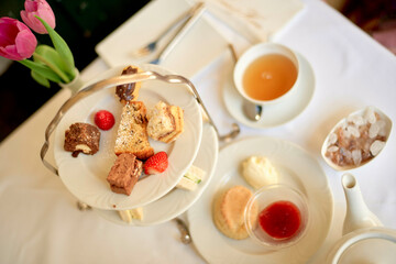 An elegant afternoon tea setup featuring a selection of cakes, a teacup, and a teapot on a table, accented by a vase with pink tulips.