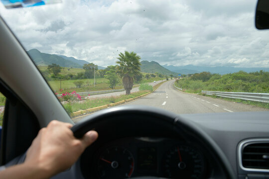 A view from the driver's perspective inside a vehicle, with the driver's left hand on the steering wheel, overlooking a curving road flanked by green landscapes under a cloudy sky.