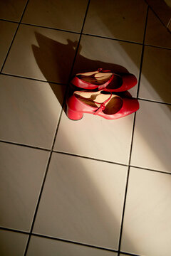 A pair of red high-heeled shoes on white tiled flooring casts a long shadow under direct sunlight.
