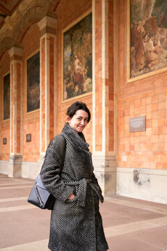 A smiling woman in a stylish houndstooth coat holding a purse stands in front of an ornate building with fresco paintings and arches.