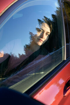 A person is looking through a car window, with their face partially obscured by reflections of trees.