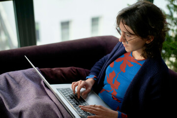 A person is sitting on a couch working on a laptop, dressed in a blue sweater with a red pattern, with a blanket over their legs.