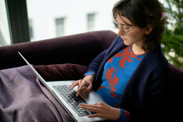 A woman is sitting on a purple couch using a laptop. She appears focused on her work, with natural light illuminating the room from a window beside her.