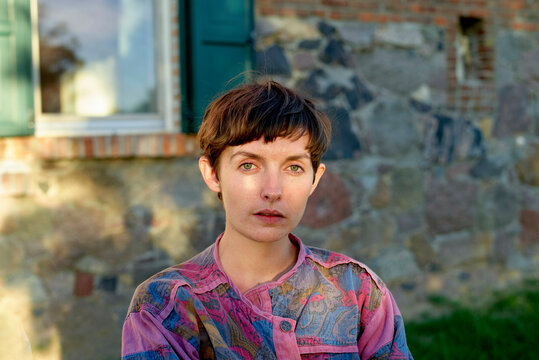 A portrait of a young woman with short hair and striking blue eyes wearing a patterned shirt, posed in front of a stone wall and window.