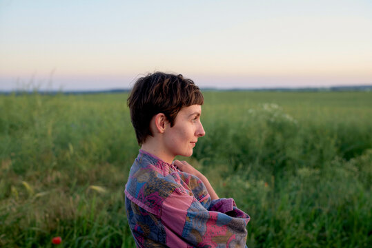 A young individual with short hair sits in a field at dusk, gazing into the distance with a serene expression.