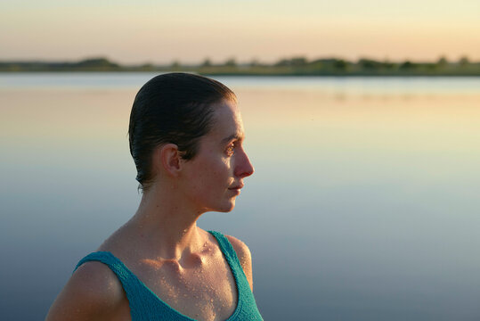 A woman is standing by a calm lake during sunset, with sunlight highlighting her profile.